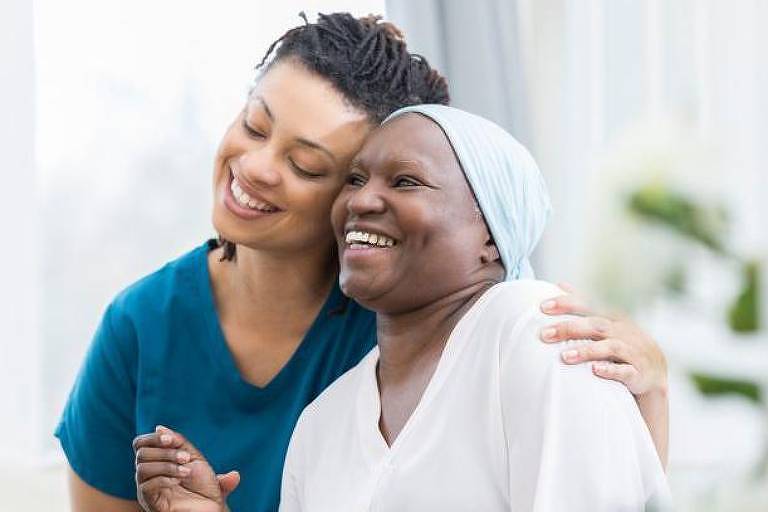 A imagem mostra uma enfermeira jovem, vestida com um uniforme azul, abraçando uma paciente idosa que está usando um lenço na cabeça e uma roupa branca. Ambas estão sorrindo