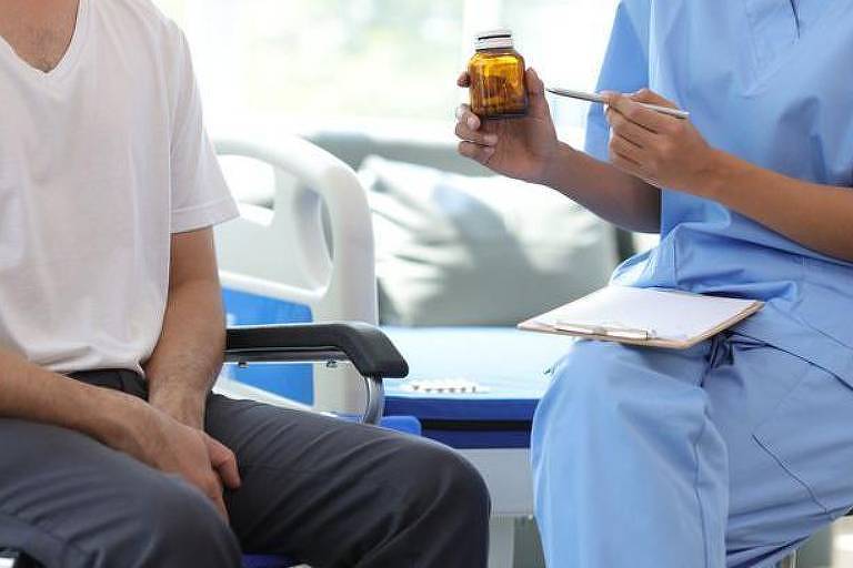 A imagem mostra, à direita, uma pessoa vestida com uniforme azul de profissional de saúde com um frasco de medicamento e uma colher, enquanto faz anotações em um bloco de papel. À esquerda, uma pessoa sentada em uma cadeira de rodas, vestindo uma camiseta branca e calça cinza, está parcialmente visível.