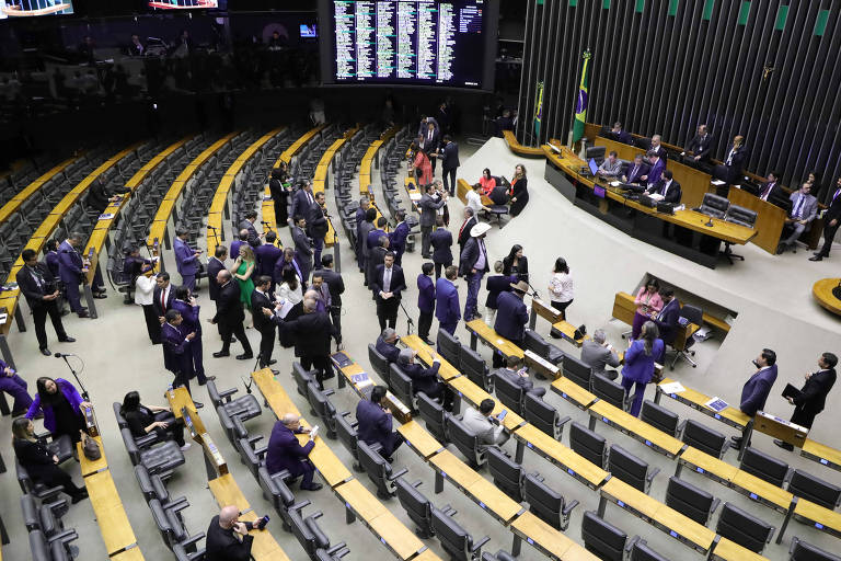 Imagem de uma sessão no plenário da Câmara dos Deputados do Brasil. A foto mostra várias pessoas, incluindo deputados e funcionários, circulando pelo espaço. As cadeiras estão dispostas em semicírculo e há um painel eletrônico ao fundo. A bandeira do Brasil está visível ao lado direito da imagem.