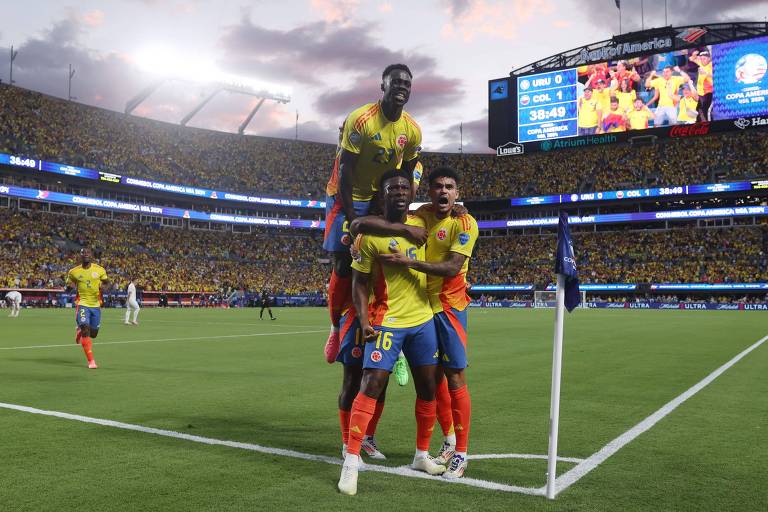 Três jogadores de futebol, vestindo uniformes amarelos e vermelhos, comemoram um gol perto da bandeira de escanteio em um estádio lotado. Um jogador está sendo levantado pelos outros dois. Ao fundo, há um placar eletrônico e uma arquibancada cheia de torcedores.
