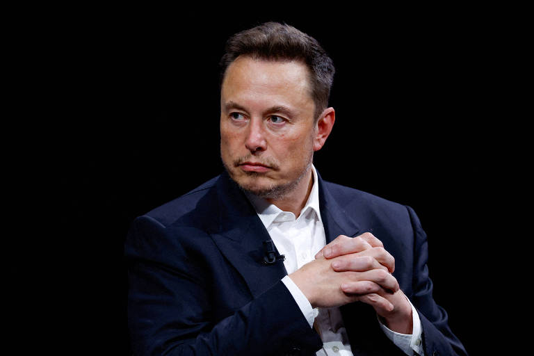 Fotografia mostra Elon Musk sentado, de mãos cruzadas na altura do peito. Ele é um homem branco, de olhos claros e cabelos castanhos. Ele olha para a direita (à esquerda da foto). Musk usa uma camisa branca e paletó azul-escuro. Fundo preto.