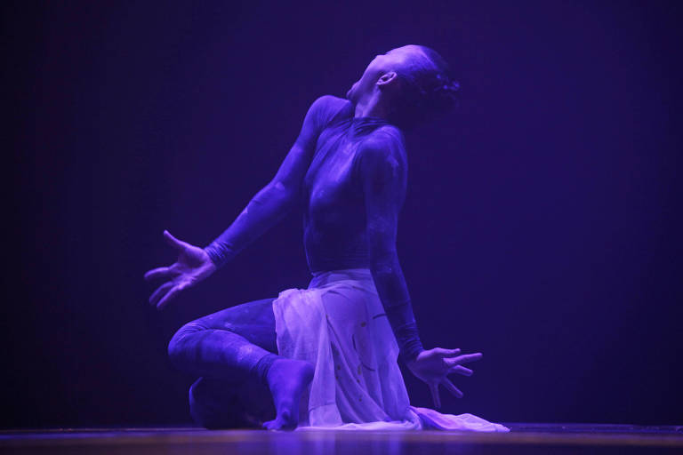 A imagem mostra uma dançarina em uma performance artística, iluminada por uma luz roxa. Ela está ajoelhada no chão, com o corpo inclinado para trás e a cabeça voltada para cima. A dançarina veste um traje justo de corpo inteiro e uma saia fluida branca. Suas mãos estão abertas e estendidas para trás, criando uma pose expressiva.