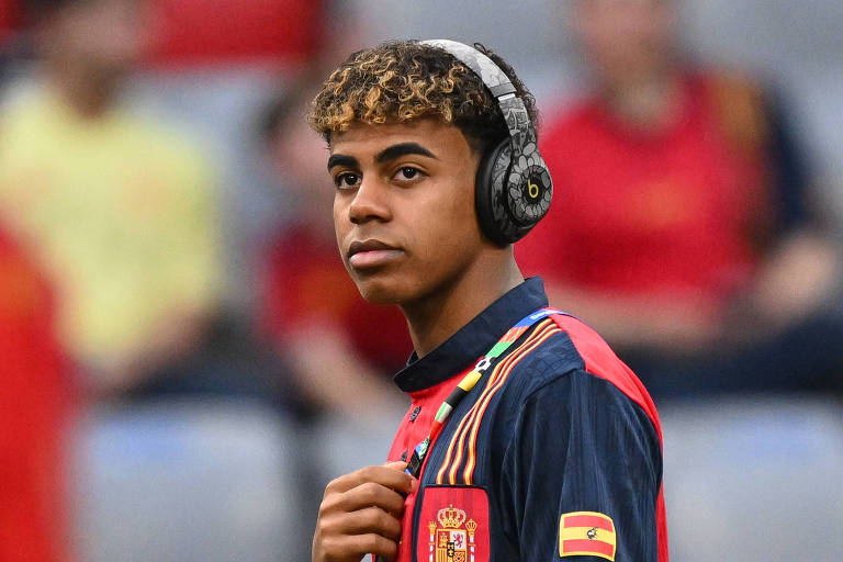 Um jovem jogador de futebol está usando uma camisa da seleção espanhola e fones de ouvido grandes. Ele tem cabelo curto e encaracolado com mechas loiras. Ao fundo, há outras pessoas desfocadas, algumas vestindo camisas vermelhas e amarelas.