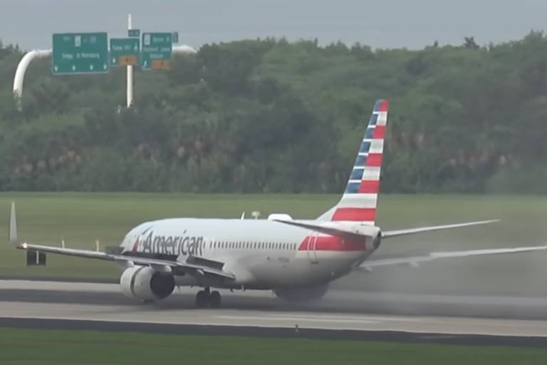 Imagem de um avião da companhia aérea American Airlines pousando em uma pista de aeroporto. O avião está em contato com o solo, com fumaça visível saindo dos pneus. Ao fundo, há uma área verde com árvores.