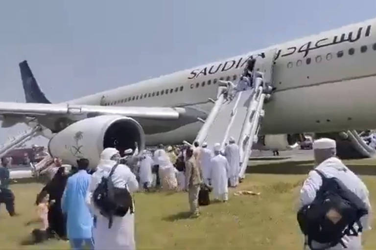 A imagem mostra um grupo de pessoas, muitas vestidas com roupas brancas tradicionais, desembarcando de um avião da Saudi Airlines. O avião está estacionado em uma área gramada, e os passageiros estão descendo por bote branco conectado à aeronave. A inscrição 'SAUDI' é visível na lateral do avião.