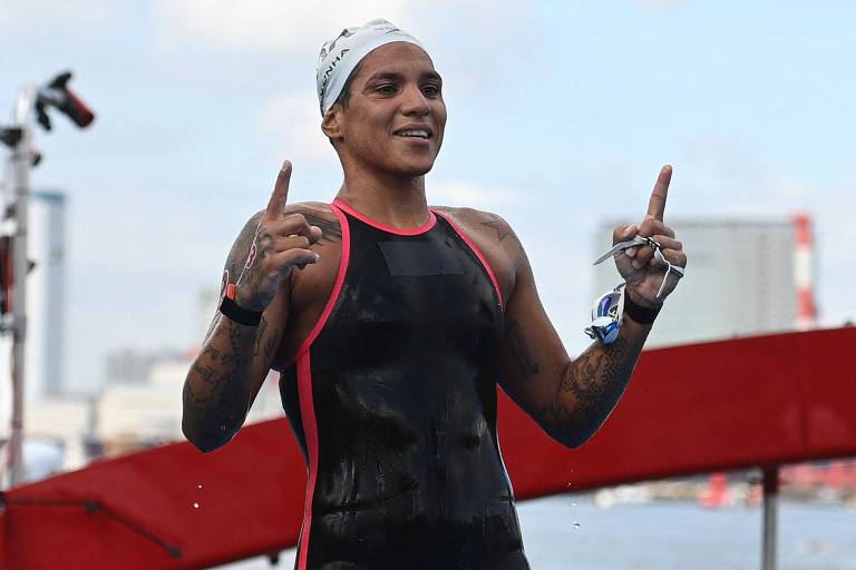 A imagem mostra uma nadadora vestindo uma roupa de natação preta com detalhes vermelhos e uma touca branca. Ela está sorrindo e levantando os dois polegares, aparentemente comemorando. Ao fundo, há uma estrutura vermelha e um céu azul.