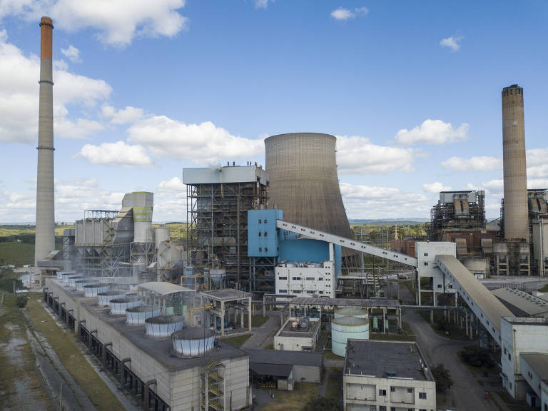 Imagem aérea de uma usina industrial com uma alta chaminé à esquerda, emitindo fumaça branca. Há várias estruturas industriais, tanques e edifícios no complexo. 
