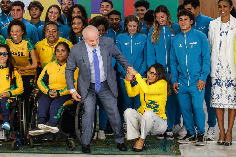 A imagem mostra um grupo de atletas paralímpicos brasileiros posando para uma foto com uma autoridade. Os atletas estão vestidos com uniformes amarelos e azuis, alguns em cadeiras de rodas. A autoridade, o presidente Lula, um homem de terno cinza e gravata azul, está ajudando a primeira-dama Janja a se levantar. Ao fundo, há uma parede colorida e mais pessoas em pé, também vestindo uniformes esportivos.