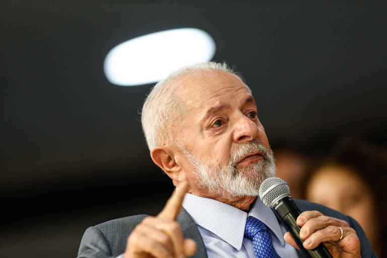 Lula aparece falando em microfone, com o dedo indicador levantado. Ele tem cabelo grisalho e usa terno.