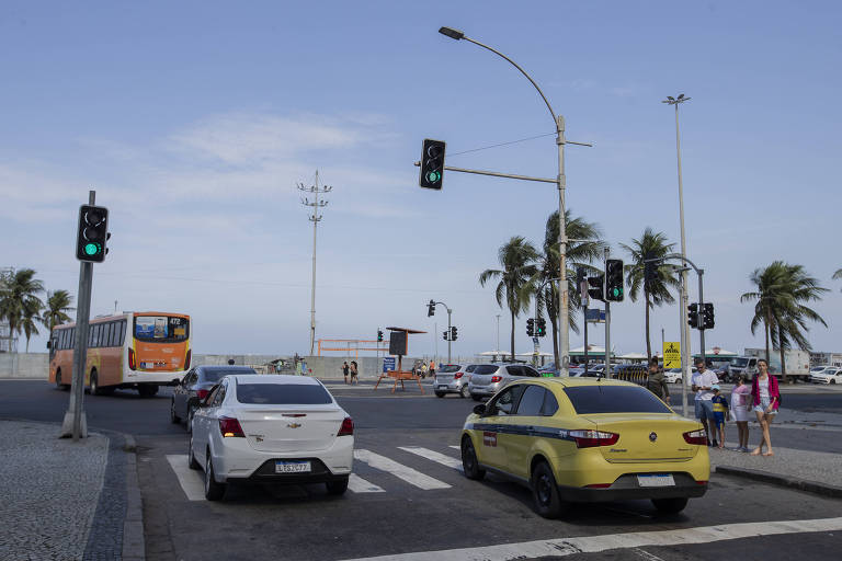 Dois veículos, um branco e outro amarelo, sendo o amarelo um táxi, trafegam pela rua. O semáforo está no sinal verde. No chão há listras brancas que indicam a faixa de pedestres. O céu está azul. O fundo da imagem é uma praia. Observa-se pequena parte da areia