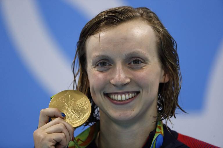 Imagem de uma nadadora sorridente segurando uma medalha de ouro. A medalha tem uma figura em relevo e está pendurada em uma fita colorida. A nadadora tem cabelo molhado e está em frente a um fundo azul.