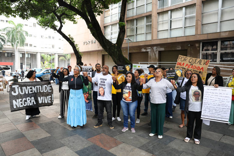 Um grupo com cerca de três dezenas de pessoas está reunido em frente ao prédio num calçadão; uma mulher segura um cartaz com a frase: "juventude negra vive"; há outros cartazes