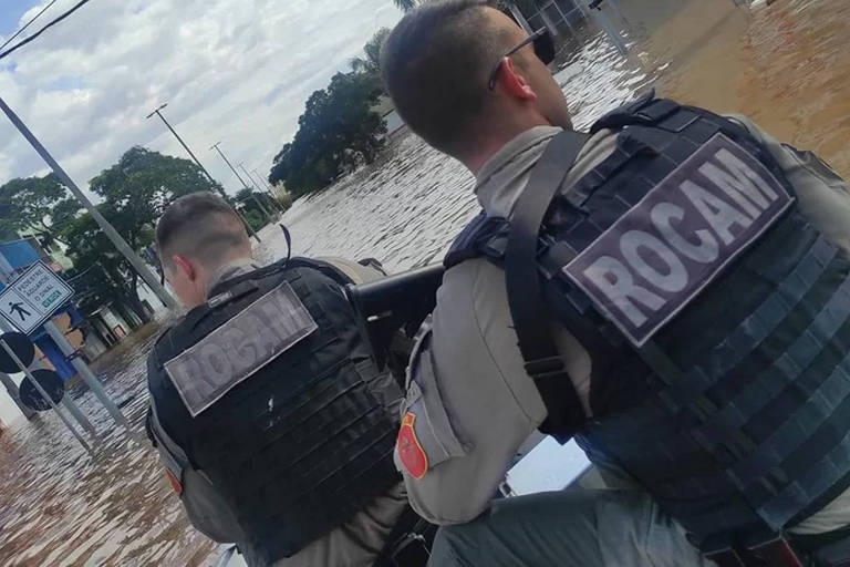A imagem mostra dois policiais de costas, vestindo coletes com a inscrição 'ROCAM'. Eles estão em uma área inundada, com água cobrindo a rua e parte das construções ao fundo. O céu está parcialmente nublado.