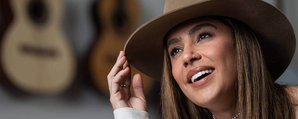 A imagem mostra uma mulher sorrindo, usando um chapéu marrom. Ela tem cabelos longos e lisos e está olhando para cima. Ao fundo, há vários violões pendurados na parede, desfocados.