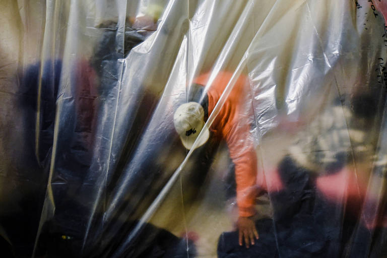 A imagem mostra uma pessoa usando um boné branco e uma camisa laranja, vista através de uma cortina de plástico transparente. A pessoa está inclinada para frente, com uma mão no chão. A imagem é desfocada, dificultando a visualização de detalhes adicionais.