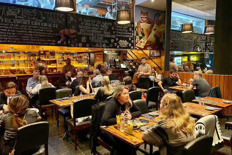 A foto mostra um salção com mesas de madeira e cadeiras pretas com clientes sentados. Ao fundo, um bar com garrafas e taças iluminadas fecham o ambiente