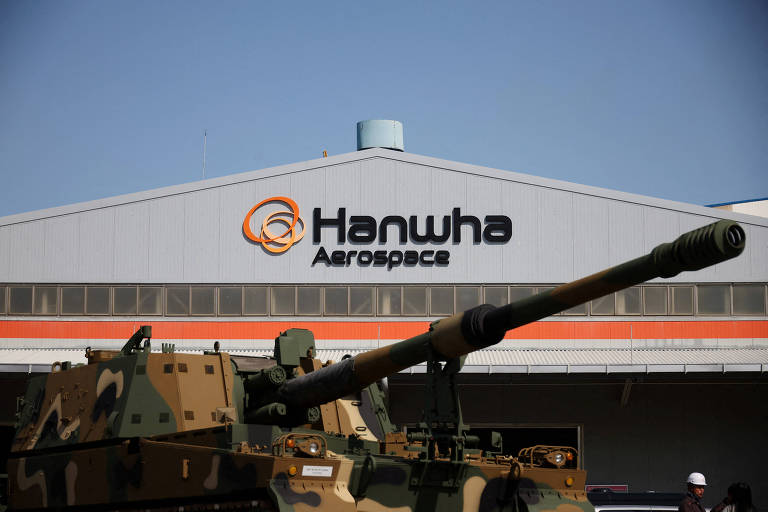 A imagem mostra a fachada de um edifício industrial com o logotipo e o nome 'Hanwha Aerospace' visíveis na parte superior. Em frente ao edifício, há um veículo militar camuflado com um grande canhão. O céu está claro e azul.