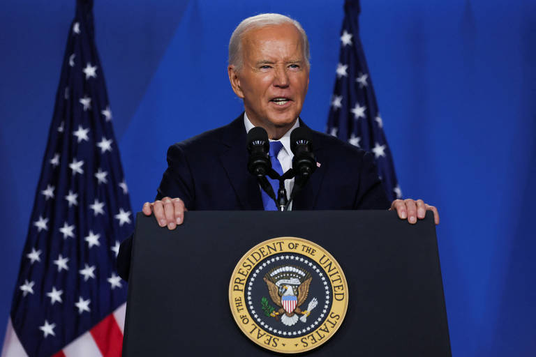 A imagem mostra uma pessoa de cabelos brancos falando em um pódio com o selo presidencial dos Estados Unidos. Atrás dele, há duas bandeiras americanas. Ele está usando um terno escuro e uma gravata azul.