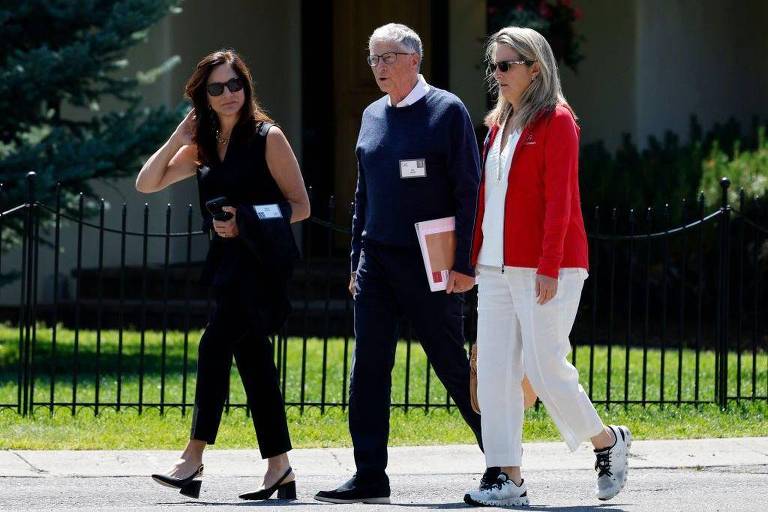 Três pessoas estão caminhando ao ar livre em um ambiente urbano. Bill Gates, cofundador da Microsoft, está no centro e vestindo um suéter azul escuro. Do lado esquerdo está uma mulher que veste roupas pretas e óculos de sol,  e segurando um caderno, e há uma outra mulher no lado direito de Bill Gates que está vestindo um casaco vermelho e calças brancas. Ao fundo, há uma cerca de ferro e vegetação.