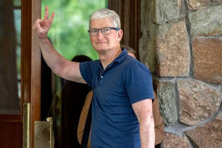 Tim Cook, diretor executivo da Apple, é um homem de óculos e cabelo grisalho está fazendo o sinal de paz com a mão direita. Ele está vestindo uma camisa polo azul e está em frente a uma porta de madeira com uma parede de pedra ao lado.
