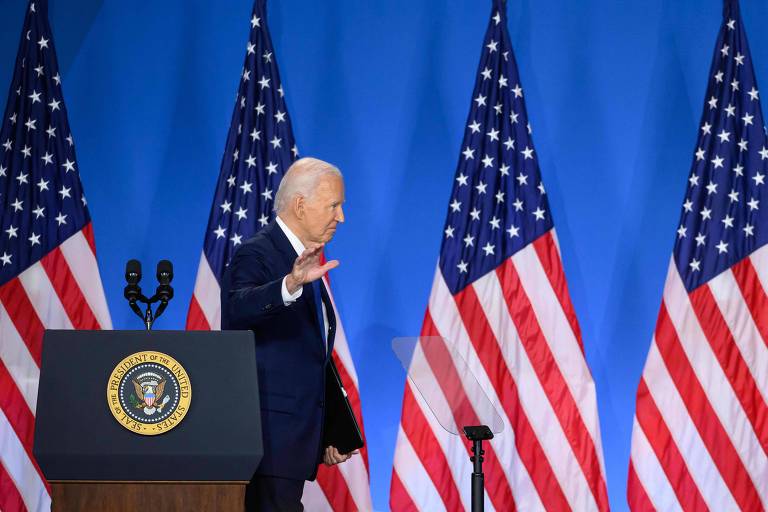 Uma pessoa de cabelos brancos, vestindo um terno escuro, está de pé em frente a um pódio com o selo presidencial dos Estados Unidos. Ele está acenando com a mão direita. Ao fundo, há quatro bandeiras dos Estados Unidos.