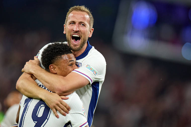 Dois jogadores de futebol estão comemorando. Um jogador está abraçando o outro, ambos vestindo uniformes brancos com detalhes em azul. O jogador que está sendo abraçado veste a camisa número 19. O fundo está desfocado, mostrando uma multidão de espectadores