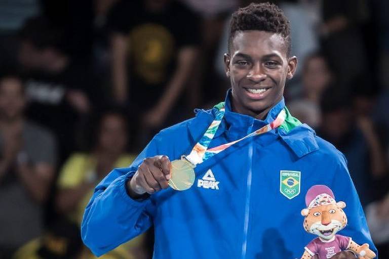 Um atleta brasileiro está sorrindo e segurando uma medalha de ouro em uma das mãos e um mascote de pelúcia na outra. Ele está vestindo um agasalho azul com o logotipo da Confederação Brasileira e a bandeira do Brasil. Ao fundo, há uma multidão desfocada.