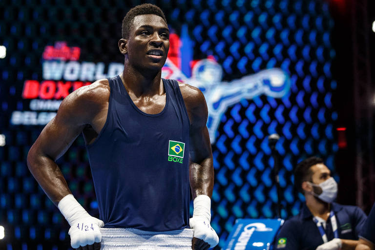 Imagem de um boxeador em um ringue de boxe, vestindo uma camiseta sem mangas azul escura com o logotipo da bandeira do Brasil. Ele está com as mãos na cintura e usa bandagens brancas nas mãos. Ao fundo, há um painel com o texto 'World Boxing' e 'Belgrade' parcialmente visíveis, além de uma figura de um boxeador em ação. Outra pessoa, usando máscara, está visível ao fundo.