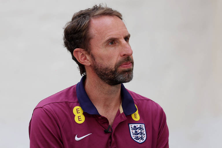 A imagem mostra um homem com barba e cabelo curto, vestindo uma camisa polo roxa com o emblema da seleção inglesa de futebol. A camisa tem detalhes em amarelo e azul escuro, e o logotipo da Nike está visível no lado direito do peito. O fundo é neutro e claro.