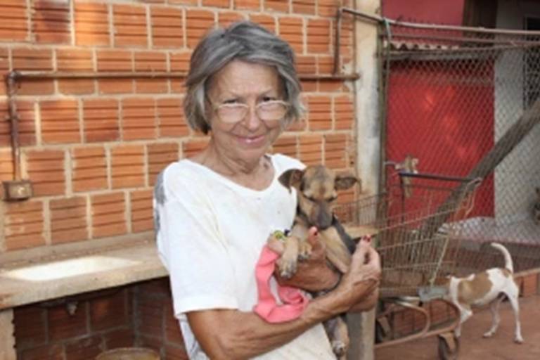 Uma mulher de cabelos grisalhos e óculos está segurando um cachorro pequeno em seus braços. Ela está vestindo uma camiseta branca. Ao fundo, há uma parede de tijolos e uma cerca de arame, com outro cachorro visível ao lado direito da imagem