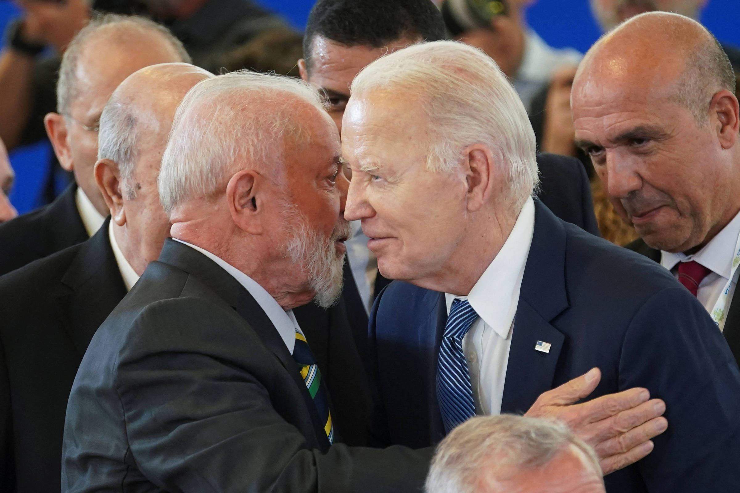 A imagem mostra dois homens se cumprimentando de forma amigável, com um deles colocando a mão no ombro do outro. Ao fundo, há várias outras pessoas, algumas com expressões de interesse. O ambiente parece ser um evento formal, com um fundo azul.