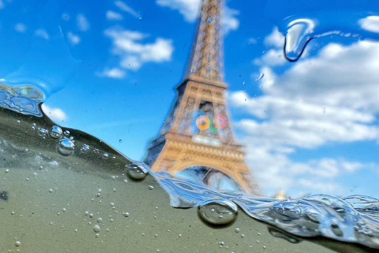A imagem mostra a Torre Eiffel em Paris, vista através de uma camada de água com bolhas. A torre está desfocada ao fundo, enquanto a água e as bolhas estão em foco na parte inferior da imagem. O céu está azul com algumas nuvens brancas.