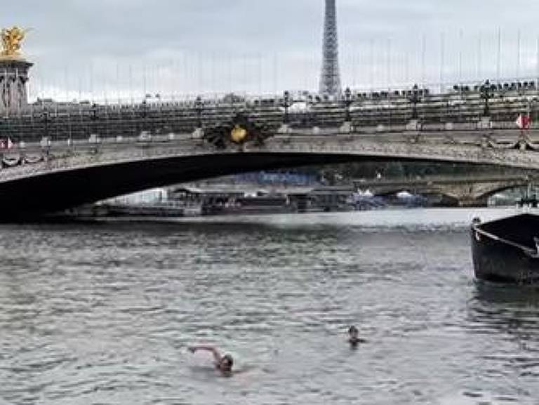 A imagem mostra uma ponte sobre o rio Sena em Paris, França, com a Torre Eiffel visível ao fundo. O céu está nublado e há algumas pessoas e veículos sobre a ponte. No rio, há duas pessoas nadando e um barco ancorado à direita.