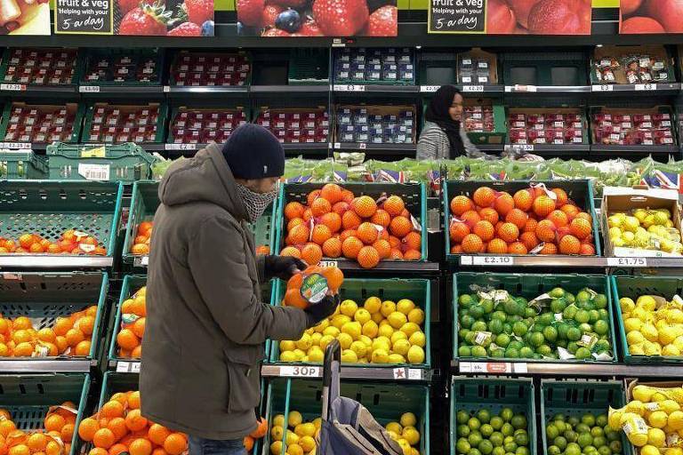 Homem está escolhendo frutas em frente a vários caixotes em um supermercado. Ele está agasalhado.