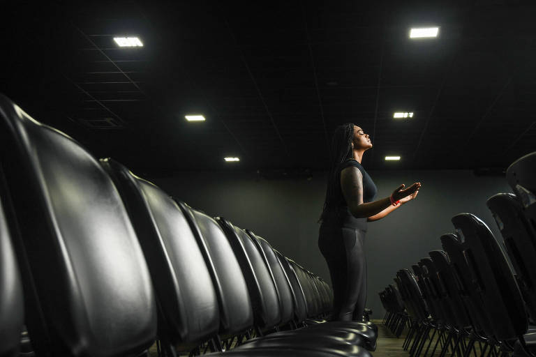 A imagem mostra uma pessoa em pé em uma sala de conferências vazia, cercada por várias fileiras de cadeiras pretas. A pessoa está de pé entre as cadeiras, com as mãos levantadas e olhando para cima. A sala é iluminada por luzes no teto.