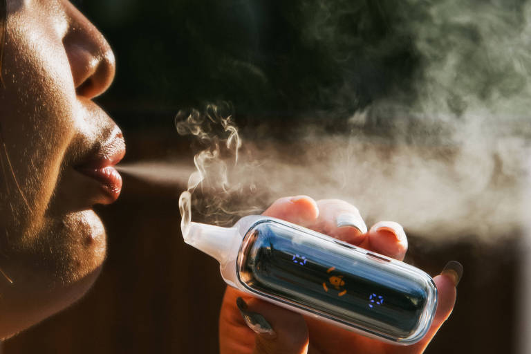 Imagem de uma pessoa soprando vapor enquanto segura um cigarro eletrônico. O vaporizador é pequeno, de cor metálica e tem uma tela com ícones iluminados. A pessoa está exalando vapor pela boca.