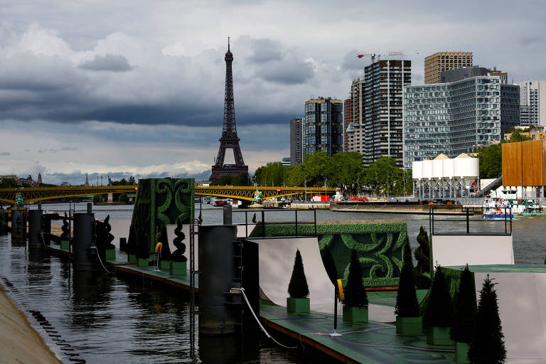 A imagem mostra uma vista do Rio Sena em Paris, com a icônica Torre Eiffel ao fundo. À direita, há vários edifícios altos e modernos. O céu está nublado, e há uma ponte amarela atravessando o rio. Na margem do rio, há estruturas decorativas verdes e pequenas árvores em vasos.