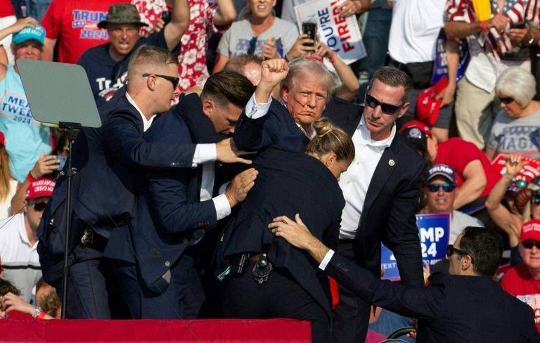Trump aparece na imagem cercado de agentes de segurança. Ele ergue o braço e tem o rosto sujo de sangue