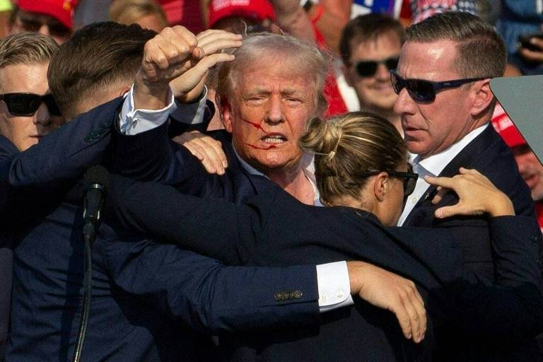 A imagem Donald Trump om expressão tensa, cercado por vários seguranças que estão o protegendo. Ele tem sangue no rosto. Os seguranças estão usando ternos escuros e óculos de sol. Ao fundo, há uma multidão de pessoas, algumas usando bonés vermelhos.