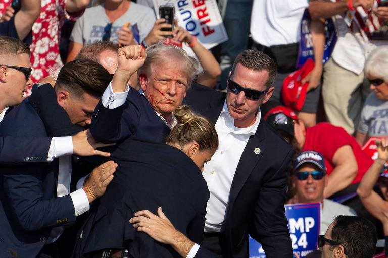 Ex-presidente Donald Trump é escoltado por seguranças após ser atingido por um tiro na orelha em comício. Com o rosto ensanguentado, ele ergue o punho esquerdo. 