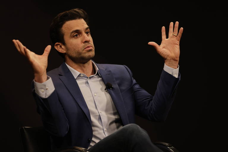 A imagem mostra um homem sentado, vestindo um terno azul e camisa branca, gesticulando com as mãos levantadas. Ele parece estar falando ou explicando algo. O fundo é escuro, destacando a figura do homem.