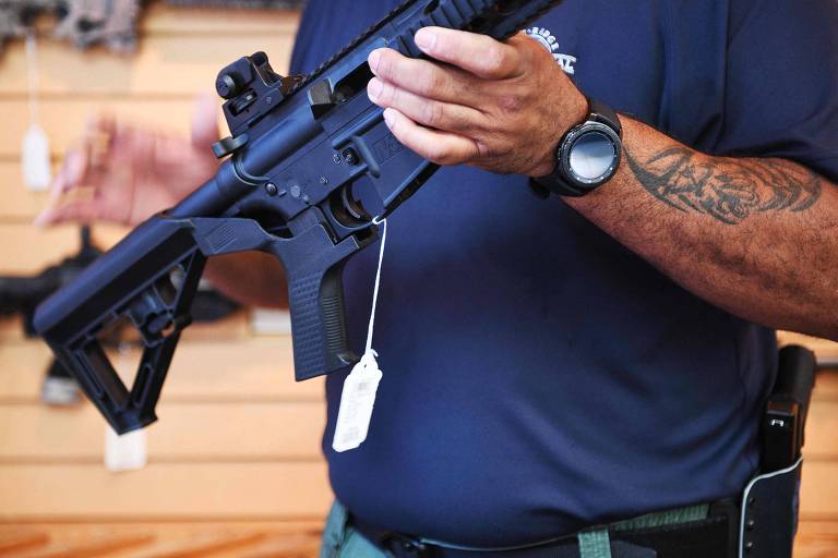 A imagem mostra uma pessoa segurando um rifle em uma loja. A pessoa está vestindo uma camisa azul escura e um relógio preto no pulso esquerdo. Há uma etiqueta branca pendurada no rifle. A pessoa também tem uma tatuagem no braço direito e um coldre preso ao cinto.