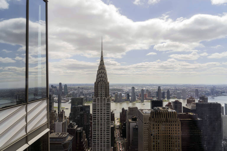 A imagem mostra uma vista panorâmica de um horizonte urbano, destacando o Edifício Chrysler no centro. O céu está parcialmente nublado com nuvens brancas e o reflexo de um prédio de vidro é visível à esquerda. Ao fundo, é possível ver outros arranha-céus e um rio.