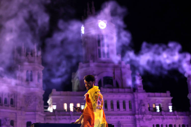 A imagem mostra uma pessoa de costas, envolta em uma bandeira da Espanha, em frente a um grande edifício iluminado com luzes roxas. Há fumaça ou névoa ao redor, criando um efeito dramático. O edifício ao fundo parece ser um marco arquitetônico, com uma torre central e um relógio visível.