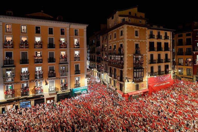Imagem de uma grande multidão vestida de vermelho e branco reunida em uma praça cercada por prédios de vários andares durante a noite. As pessoas estão celebrando, e muitas estão nas varandas dos prédios. Há uma grande faixa vermelha visível à direita da imagem.