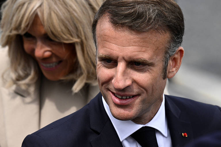 Imagem de Emmanuel Macron, presidente da França, que é um homem sorrindo, vestindo um terno preto e gravata preta, com uma mulher ao fundo desfocada, também sorrindo. O homem tem cabelo curto e castanho, enquanto a mulher tem cabelo loiro e veste um casaco claro.