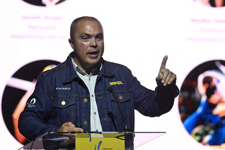 A imagem mostra um homem careca usando uma jaqueta jeans, falando em um púlpito com um microfone. Ele está gesticulando com a mão esquerda levantada.
