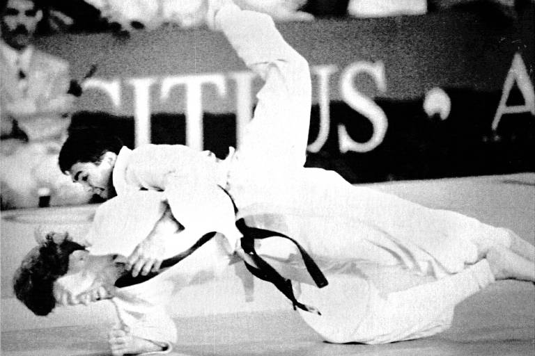 A imagem em preto e branco mostra dois judocas em ação durante uma luta. Um dos judocas está no ar, sendo arremessado pelo outro, que está no tatame. Ambos estão vestindo quimonos brancos e faixas pretas.