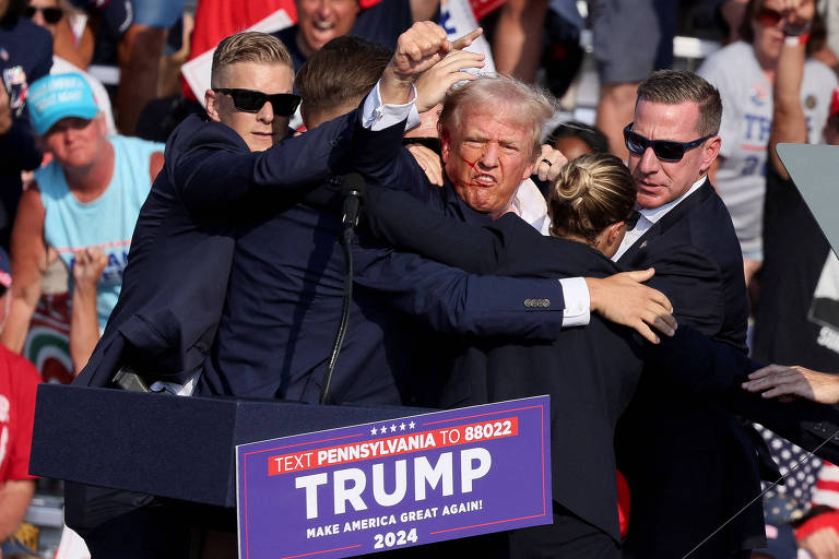 A imagem mostra um comício político com um grupo de seguranças ao redor de Donald Trump, que tem o rosto ensanguentado e está sendo protegido. Há uma multidão ao fundo, algumas pessoas usando bonés e segurando cartazes.