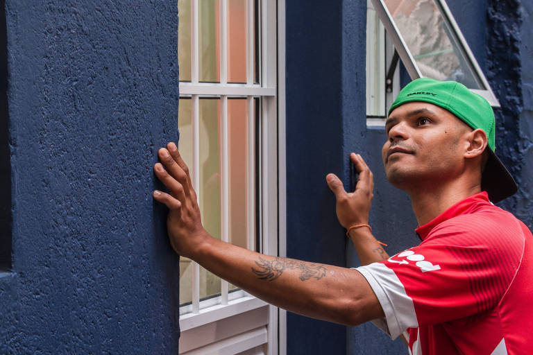 Um homem usando boné verde e camiseta vermelha está observando uma janela com grade branca em uma parede azul. Acima da janela, há uma placa com o texto 'CASACOR'. O homem está com as mãos apoiadas na parede e na janela, olhando para cima.